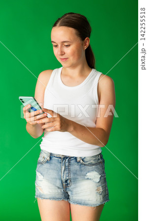 Portrait of european teen girl dressed in stylish shorts scrolling smartphone standing against green screen Portrait of european teen girl dressed in stylish shorts scrolling smartphone standing against green screen 142255518