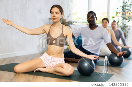 Woman in activewear sitting on yoga mat during group workout with fitness ball at gym. Healthy lifestyle concept Woman in activewear sitting on yoga mat during group workout with fitness ball at gym. Healthy lifestyle concept 142255553