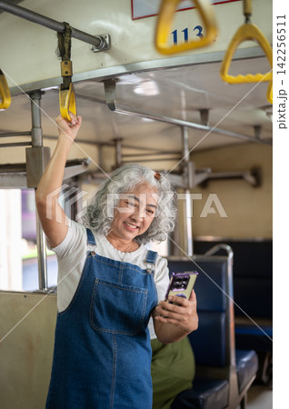 Asian senior woman smiling and looking at phone while standing holding handrail in a moving train. Asian senior woman smiling and looking at phone while standing holding handrail in a moving train. 142256511