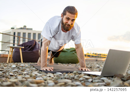 A bearded man with his hair in a bun performs a plank exercise on a yoga mat on a rooftop terrace while looking at a laptop 142257095