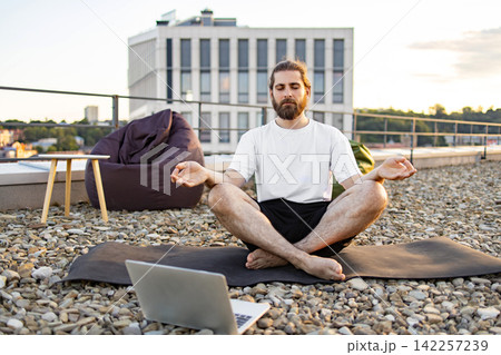 A man with a beard practices yoga and meditation on a rooftop terrace, using a laptop for guidance while sitting on a mat surrounded by pebbles 142257239