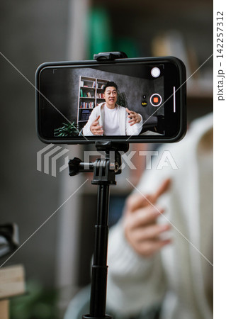 A man is actively gesturing while recording himself on a smartphone mounted on a tripod, creating content for his audience A man is actively gesturing while recording himself on a smartphone mounted on a tripod, creating content for his audience 142257312