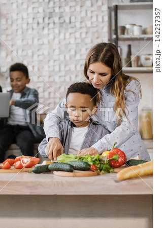 A mother and her two sons are preparing a healthy meal together in a bright, modern kitchen, chopping fresh vegetables for a salad A mother and her two sons are preparing a healthy meal together in a bright, modern kitchen, chopping fresh vegetables for a salad 142257357