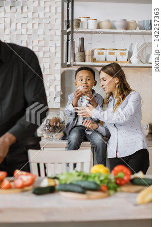 A mother and son are in the kitchen, with a father preparing food in the foreground, creating a warm family moment A mother and son are in the kitchen, with a father preparing food in the foreground, creating a warm family moment 142257363