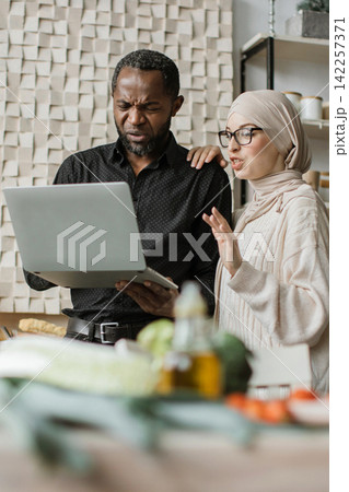 A man and woman in a kitchen look at a laptop, possibly reviewing a recipe or discussing meal preparation 142257371