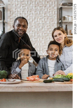 A diverse family with parents and two sons smiling while preparing healthy food in a modern kitchen, showcasing togetherness and healthy eating A diverse family with parents and two sons smiling while preparing healthy food in a modern kitchen, showcasing togetherness and healthy eating 142257375