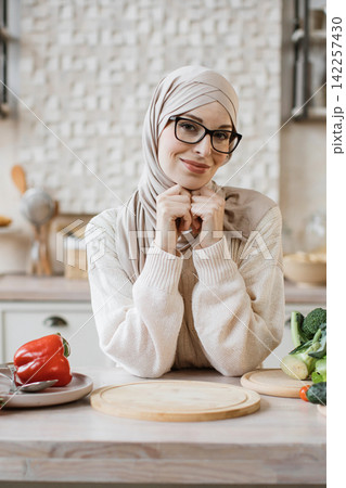 A smiling woman wearing a hijab and glasses is in a kitchen, ready to prepare a healthy meal with fresh vegetables 142257430