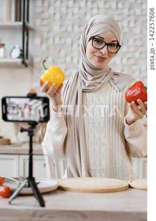 A woman wearing a hijab and glasses records a cooking tutorial on her smartphone, holding colorful bell peppers A woman wearing a hijab and glasses records a cooking tutorial on her smartphone, holding colorful bell peppers 142257606