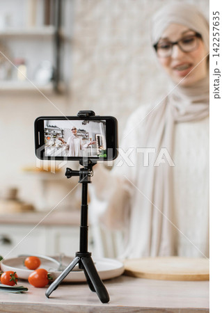 A woman wearing a hijab and glasses records a cooking tutorial on her smartphone placed on a tripod in a bright kitchen setting A woman wearing a hijab and glasses records a cooking tutorial on her smartphone placed on a tripod in a bright kitchen setting 142257635