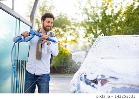 A smiling caucasian man is actively washing his car using a high-pressure water jet and foam at an outdoor car wash station 142258533