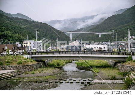 曇り空の下で山靄が漂う緑豊かな山並みと小川にかかる橋と街並みの風景 曇り空の下で山靄が漂う緑豊かな山並みと小川にかかる橋と街並みの風景 142258544