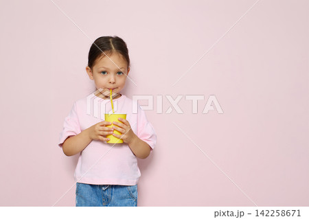 Young girl drinking a refreshing beverage from a yellow paper cup with a straw against a pink background, expressing childhood and hydration Young girl drinking a refreshing beverage from a yellow paper cup with a straw against a pink background, expressing childhood and hydration 142258671