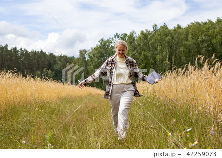 Woman walks along grassy path between ripe wheat fields carrying digital tablet during field inspection in summer. Concept of agricultural consulting, field research and sustainable crop management. 142259073
