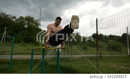 Strong man trains abdominal muscles raising legs on parallel bars at sports ground. Young shirtless sportsman exercising at nature. Sporty athlete working out at nature. Concept of sportive lifestyle Strong man trains abdominal muscles raising legs on parallel bars at sports ground. Young shirtless sportsman exercising at nature. Sporty athlete working out at nature. Concept of sportive lifestyle 142260827