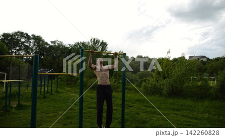 Muscular athlete doing pull ups on horizontal bar at sports ground. Young shirtless sportsman exercising at nature. Strong sporty man training outdoor. Concept of healthy and active lifestyle. Slow mo Muscular athlete doing pull ups on horizontal bar at sports ground. Young shirtless sportsman exercising at nature. Strong sporty man training outdoor. Concept of healthy and active lifestyle. Slow mo 142260828
