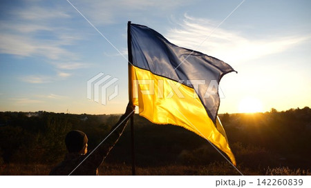 Young soldier of ukrainian army lifted blue-yellow banner in honor of the victory against russian aggression at sunset. Male military in uniform raised a waving flag of Ukraine at countryside 142260839