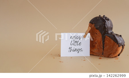 ENJOY LITTLE THINGS saying caption on paper note Heart shaped dark chocolate croissant on beige background with copy space. Puff pastry new trendy sweet dessert bakery. Creamy dough 142262999