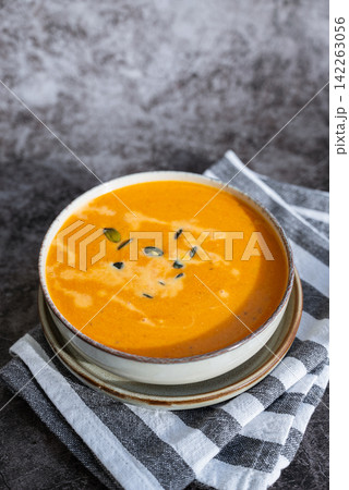 Bowl of creamy pumpkin soup topped with pumpkin seeds. Rich orange puree is served in rustic bowl and decorated with crunchy seeds creating an appetizing autumn meal presentation. Concept of seasonal Bowl of creamy pumpkin soup topped with pumpkin seeds. Rich orange puree is served in rustic bowl and decorated with crunchy seeds creating an appetizing autumn meal presentation. Concept of seasonal 142263056