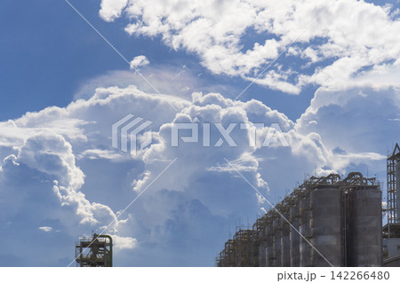 Rows of cylindrical storage tanks and pipe structures at a petrochemical plant silhouetted against a dramatic blue sky with towering cumulonimbus clouds creating a striking contrast between nature and 142266480