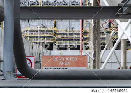 A restricted area warning sign reading DO NOT ENTER at a petrochemical industrial plant with yellow railings, steel structures and large pipelines visible in the background. A restricted area warning sign reading DO NOT ENTER at a petrochemical industrial plant with yellow railings, steel structures and large pipelines visible in the background. 142266481