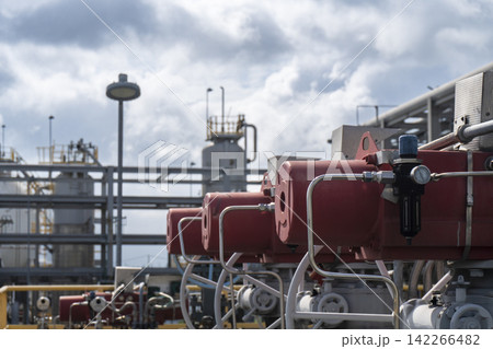 A row of red valve actuators with pressure gauges and connected piping lined up at a petrochemical plant facility under a cloudy sky showing automated flow control equipment. 142266482