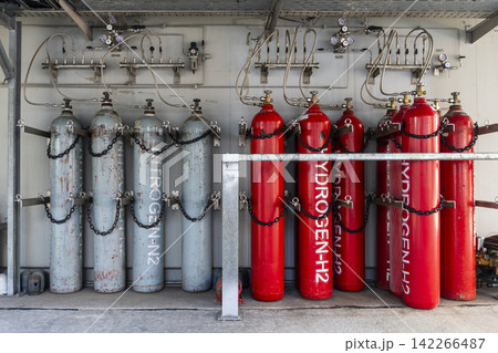 Industrial gas storage facility with red hydrogen H2 cylinders and gray nitrogen N2 cylinders chained in rows connected to pressure gauges and piping manifolds on a wall panel. Industrial gas storage facility with red hydrogen H2 cylinders and gray nitrogen N2 cylinders chained in rows connected to pressure gauges and piping manifolds on a wall panel. 142266487