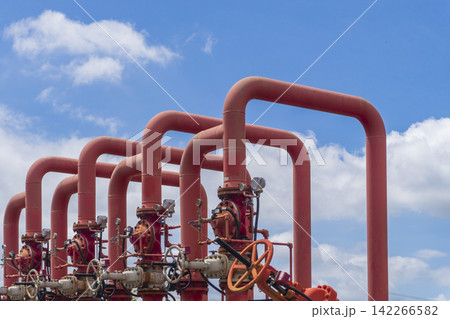 Rows of red fire protection pipelines and valve manifolds with pressure gauges and handwheel valves aligned against a bright blue sky at an industrial plant forming a bold geometric composition. Rows of red fire protection pipelines and valve manifolds with pressure gauges and handwheel valves aligned against a bright blue sky at an industrial plant forming a bold geometric composition. 142266582