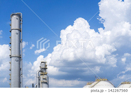 Tall distillation columns and a spherical gas storage tank at a petrochemical plant set against a vivid blue sky with dramatic white cumulus clouds creating a striking contrast. Tall distillation columns and a spherical gas storage tank at a petrochemical plant set against a vivid blue sky with dramatic white cumulus clouds creating a striking contrast. 142267560