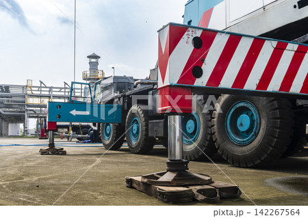 A large mobile crane with red striped body and blue wheels deployed with outriggers extended at a petrochemical plant site in an industrial heavy equipment close-up shot. 142267564