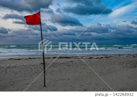 Red warning flag standing tall on a beach with cloudy skies and waves Red warning flag standing tall on a beach with cloudy skies and waves 142268932