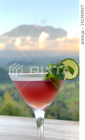 Red cocktail in martini glass garnished with lime slice and mint, set on outdoor table with mountain landscape, soft pastel sky, bright refreshing drink for summer leisure Red cocktail in martini glass garnished with lime slice and mint, set on outdoor table with mountain landscape, soft pastel sky, bright refreshing drink for summer leisure 142269927