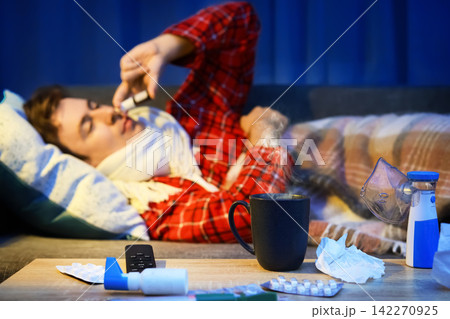 Sick man with cold and flu symptoms uses nasal spray on a couch. Home treatment for respiratory infection includes medicine, hot drink and selfcare for illness recovery. 142270925