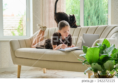 10-year-old girl studying on sofa with laptop and notebook near black cat 10-year-old girl studying on sofa with laptop and notebook near black cat 142272115