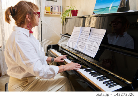 Woman practicing piano at home, reading sheet music and playing an upright piano during a private piano-class session, focused expression, casual interior and musical practice. 142272326