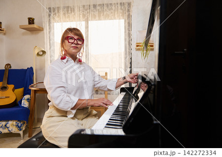 Woman seated at a piano in a sunlit living room, playing and adjusting the instrument. Warm, relaxed scene with guitar, books and plant suggesting home music practice, teaching and creativity. Woman seated at a piano in a sunlit living room, playing and adjusting the instrument. Warm, relaxed scene with guitar, books and plant suggesting home music practice, teaching and creativity. 142272334