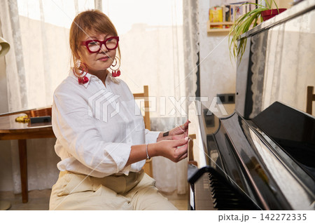 Middle-aged woman in red glasses tuning a glossy grand piano at home during a casual piano class, showing focus, patience and musical dedication in a cozy living-room setting. 142272335