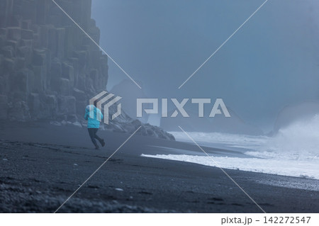 Runner in blue jacket on black sand beach Iceland with misty basalt cliffs Runner in blue jacket on black sand beach Iceland with misty basalt cliffs 142272547
