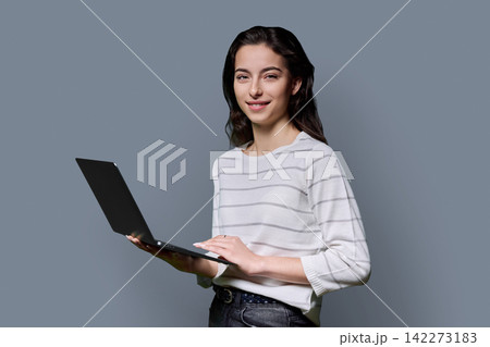 Young female student holding laptop computer and looking at camera on grey background 142273183