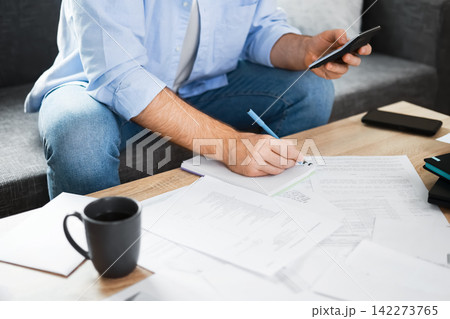 Man calculating household expenses and utility bills at home. He faces financial pressure from inflation and economic crisis, writing a budget to manage rising heating costs. Man calculating household expenses and utility bills at home. He faces financial pressure from inflation and economic crisis, writing a budget to manage rising heating costs. 142273765