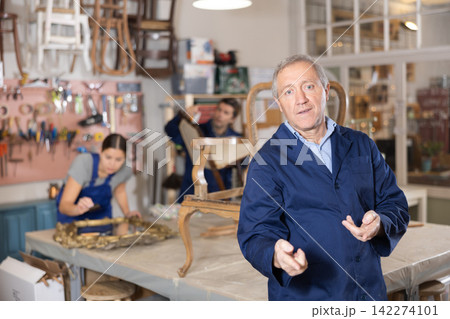 Professional elderly craftsman restorer in uniform looking at camera while working in repair shop Professional elderly craftsman restorer in uniform looking at camera while working in repair shop 142274101