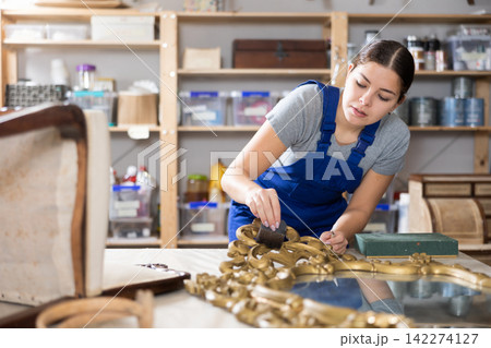 Girl restorer carefully covers carved wooden frame of antique wall mirror with thin layer of varnish Girl restorer carefully covers carved wooden frame of antique wall mirror with thin layer of varnish 142274127