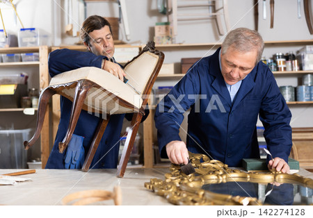 Senior male furniture workshop worker and his apprentice repairing vintage mirror and chair Senior male furniture workshop worker and his apprentice repairing vintage mirror and chair 142274128