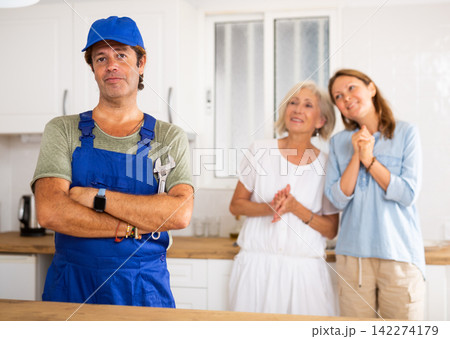 Portrait of successful plumber in kitchen. Happy apartment owners in background Portrait of successful plumber in kitchen. Happy apartment owners in background 142274179