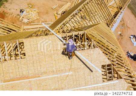 Worker stands on roof uses tools to attach materials while construction continues on new house nearby. 142274712