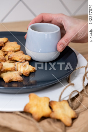 Crispy pastry. Homemade biscuits. Female hand holding beverage cup on served star cookies plate. Crunchy confectionery treat tasting time. Crispy pastry. Homemade biscuits. Female hand holding beverage cup on served star cookies plate. Crunchy confectionery treat tasting time. 142277074
