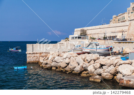 Colorful fishing boats moored by a rocky breakwater and stone quay. Valletta, Malta Colorful fishing boats moored by a rocky breakwater and stone quay. Valletta, Malta 142279468