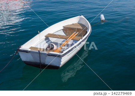 A small white rowboat moored to a buoy and floating on calm blue harbor water A small white rowboat moored to a buoy and floating on calm blue harbor water 142279469