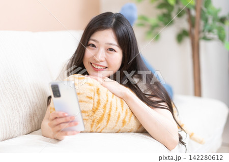 ソファで寝そべってスマホを持って微笑む若い女性 142280612