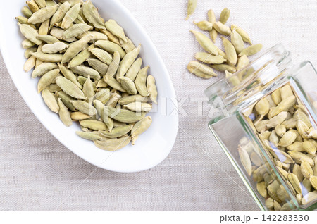 Whole, dried cardamom pods, in oval white bowl on linen. Sometimes called cardamon, or cardamum, greenish fruits with seeds of Elettaria cardamomum, a spice with sharp, strong and punchy aroma. Photo Whole, dried cardamom pods, in oval white bowl on linen. Sometimes called cardamon, or cardamum, greenish fruits with seeds of Elettaria cardamomum, a spice with sharp, strong and punchy aroma. Photo 142283330