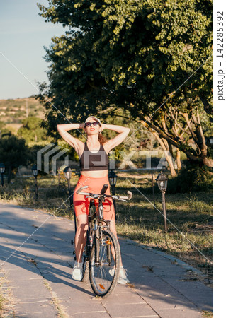 Relaxing park break enjoyed by fit young woman resting with hands behind head next to her bicycle during morning sunshine Relaxing park break enjoyed by fit young woman resting with hands behind head next to her bicycle during morning sunshine 142285392
